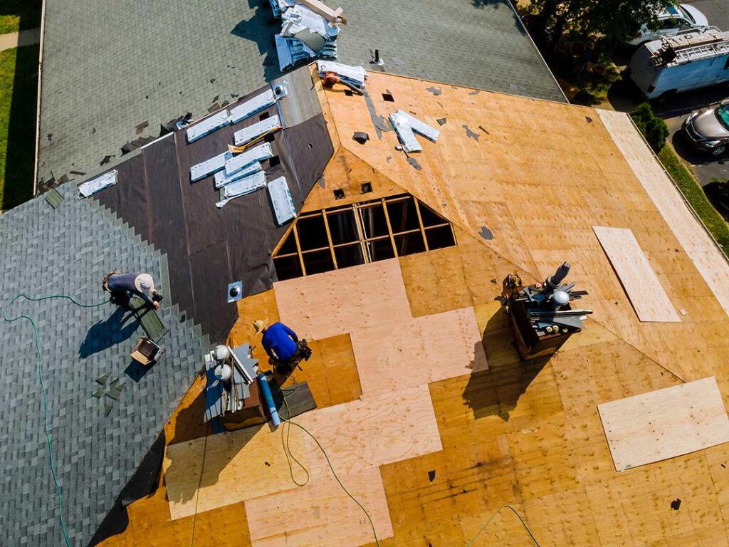 Inspector examines shingles and flashing beside a ladder while another person checks the attic.